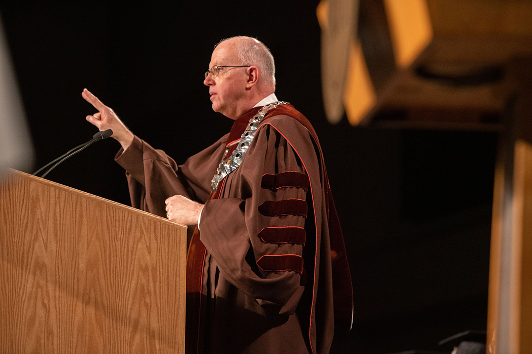 RIT president David Munson wearing graduation regalia and speaking at a podium.