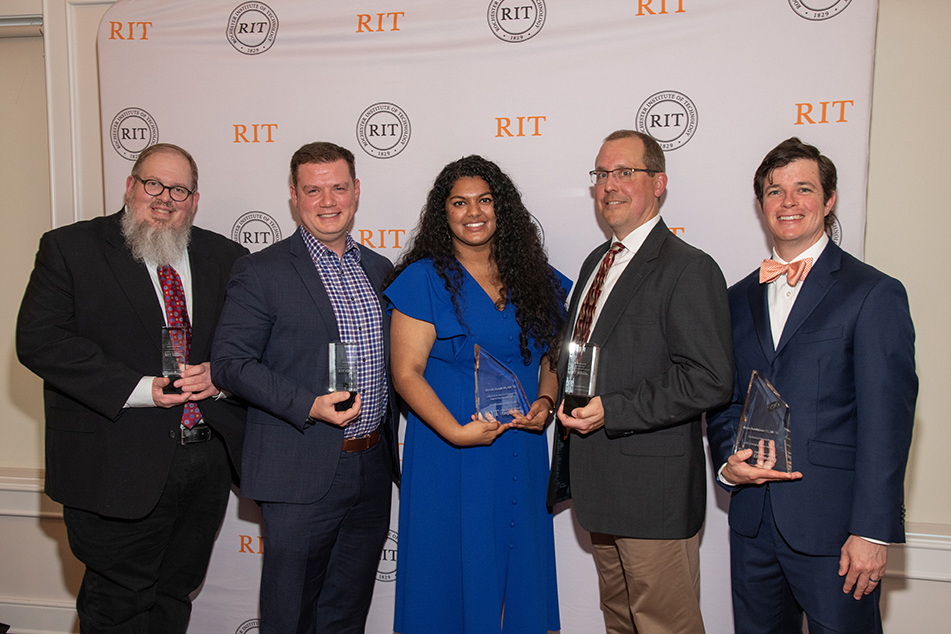 Five people standing in front of a logo backdrop, smiling and holding awards.