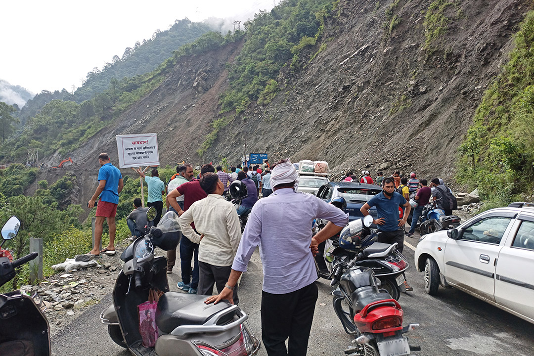 people standing outside their cars and scooters on a road in a hill that's been covered by a landslide.