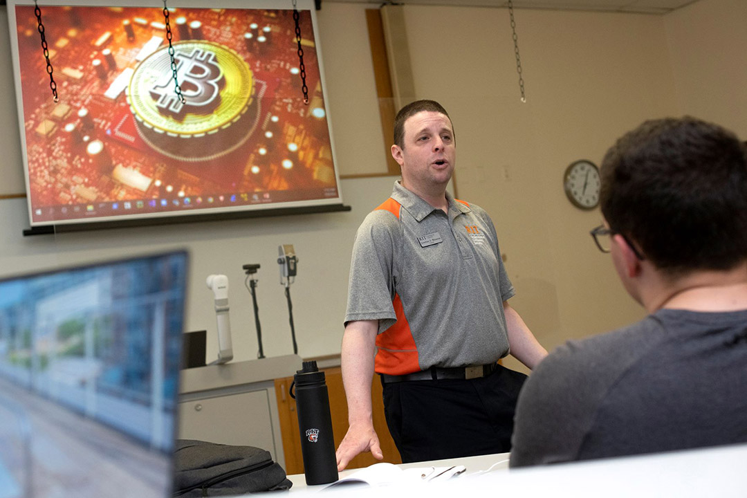 Faculty member in front of screen with image of cryptocurrency.