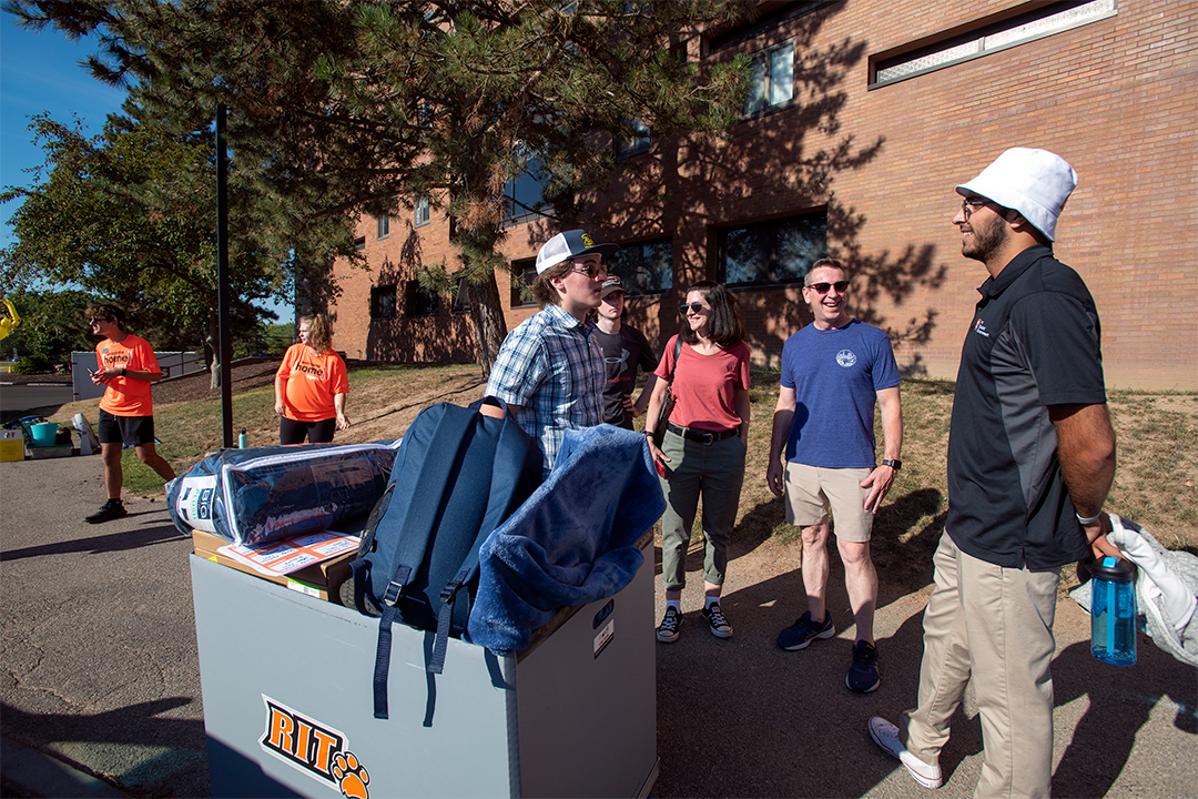 A small group of parents and college students speak outside of a building in front of a large bin filled with luggage.