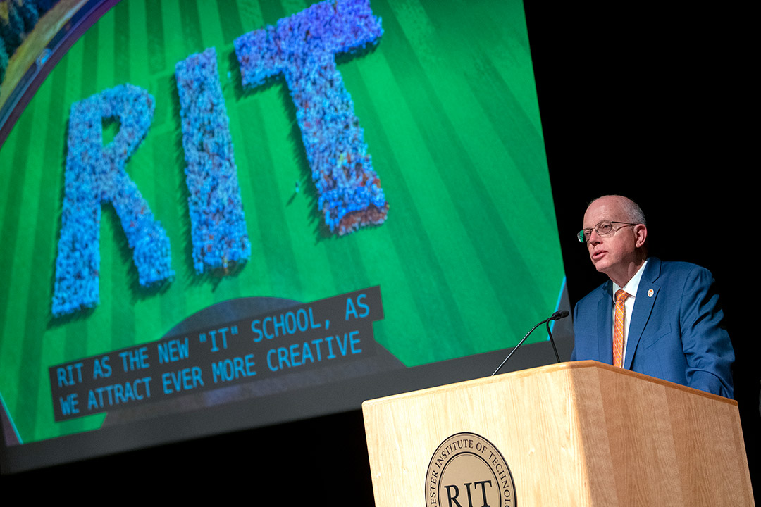 RIT President Munson speaking at podium with a power point projection in the background.