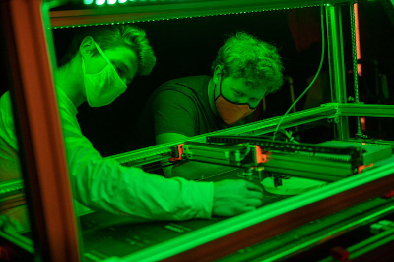 green overlay on image of two students working on a 3D printer.