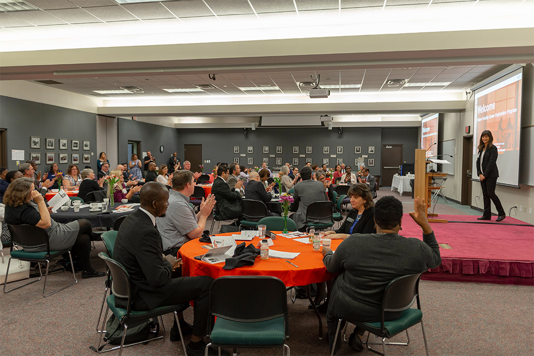 people sitting at round tables applauding a presenter on a small stage.