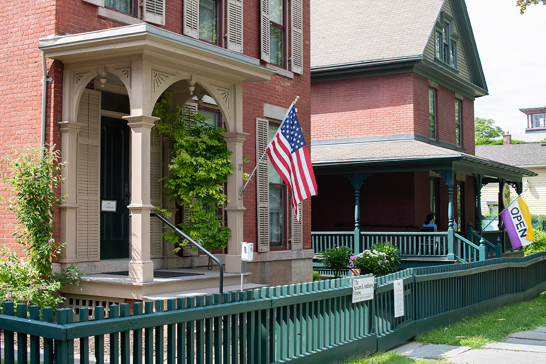 a small brick house with an American flag and surrounded by a green picket fence.
