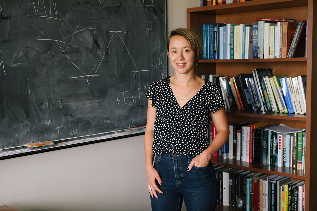 woman standing in a classroom next to a chalkboard and bookcase.