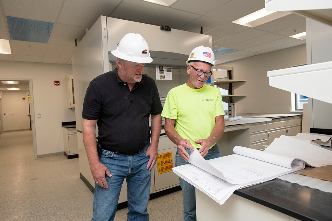 two people in a lab wearing hard hats looking at blueprints.