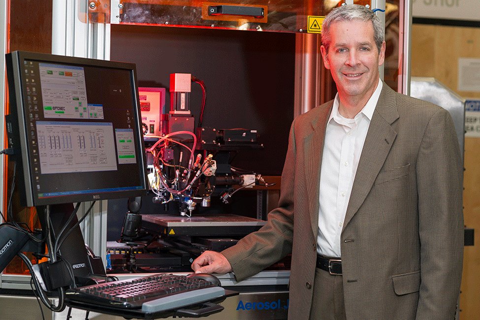 Professor Denis Cormier standing in front of a 3D printer with a computer screen and keyboard to his right.