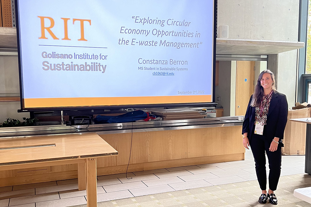 person standing next to a large screen with a projection showing the RIT Golisano Institute for Sustainability logo.