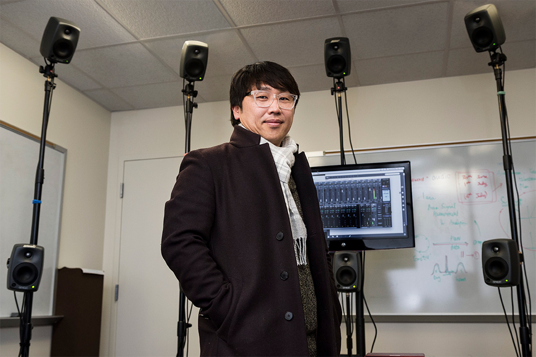 A man wearing glasses and a peacoat stands in front of eight small speakers and a digital audio control board.