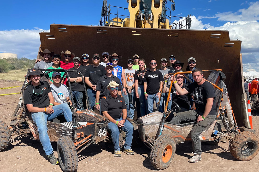 team of people and two off-road Baja cars in the bucket of a front loader.