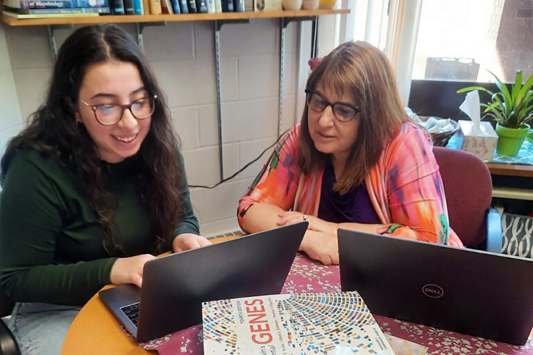 professor and student sitting at a table working on laptops.