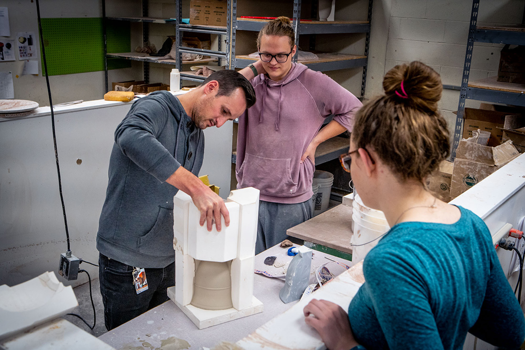 two students watching as a professor unmolds an upside down clay vase.