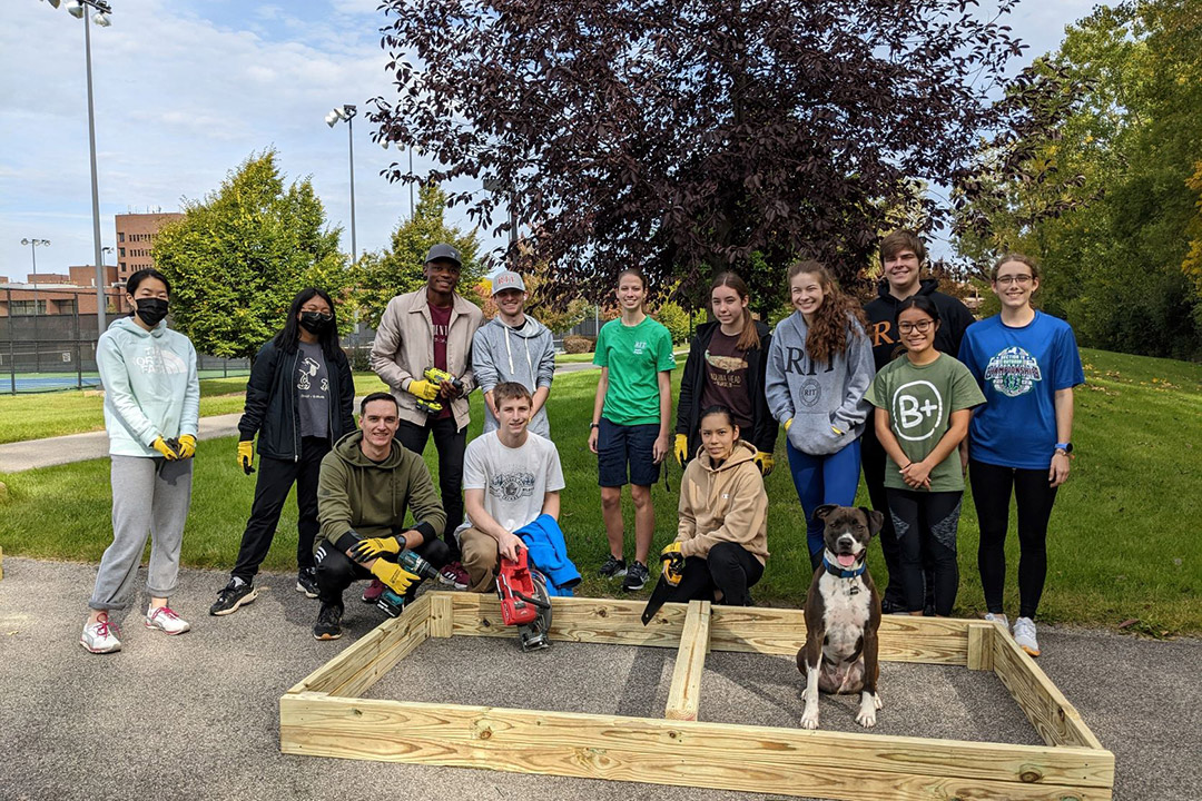 13 people and a dog posing around and in a frame for a raised garden bed.