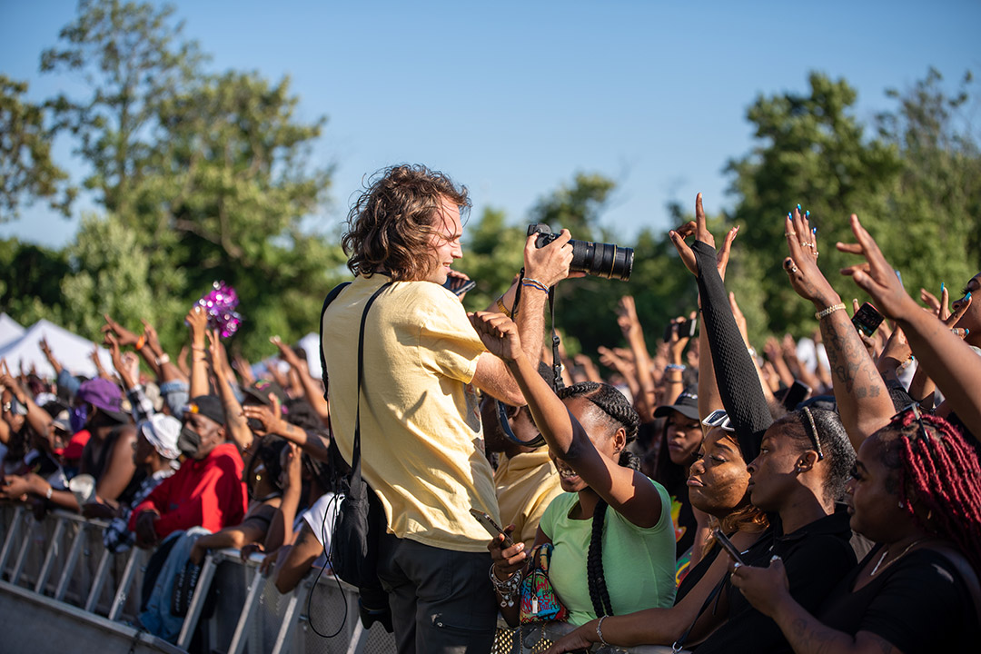 photographer with camera facing a crowd of people with the arms raised in the air.