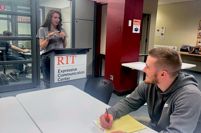 student sitting at a table taking notes while another student speaks at a podium.