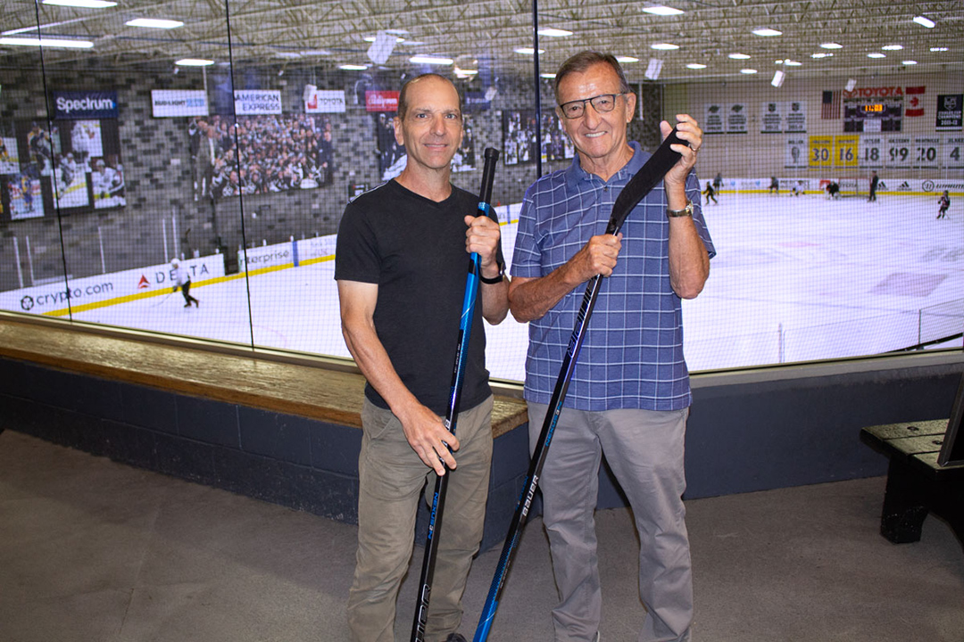 father and son holding hockey sticks posing outside of the ice of a hockey arena.
