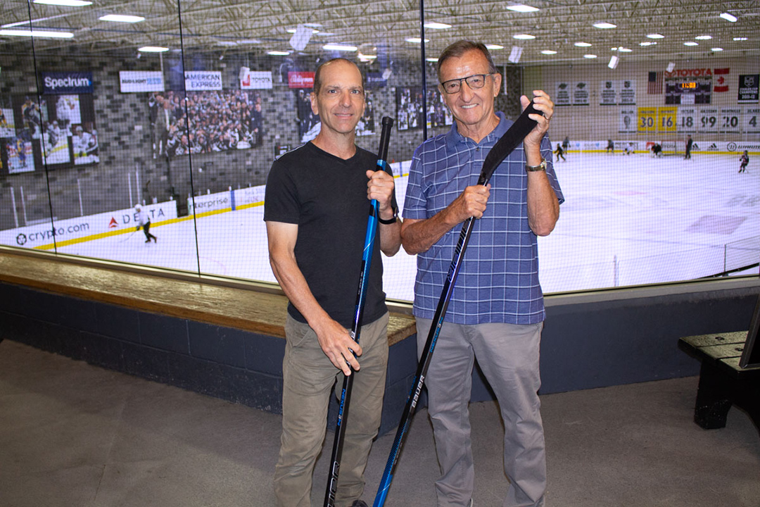 two men standing just outside an ice rink holding hockey sticks.