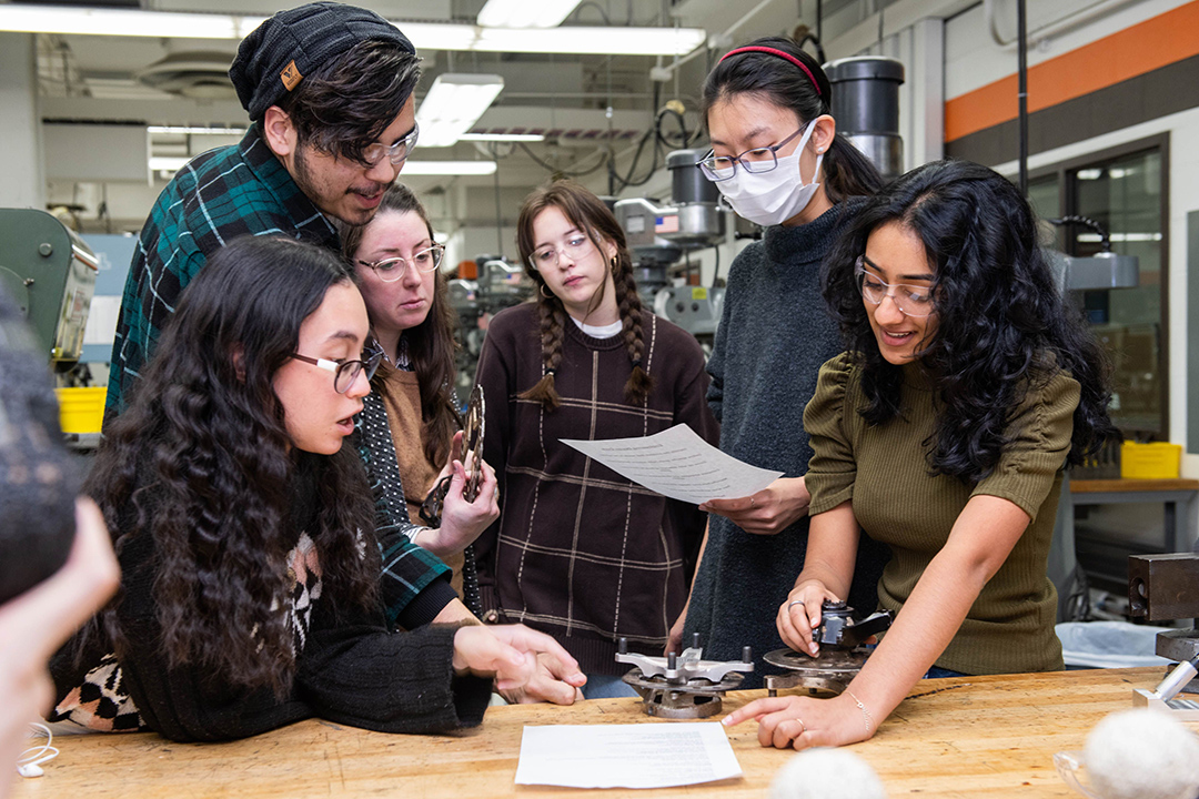 six students crowded around a table looking at a paper and car parts.