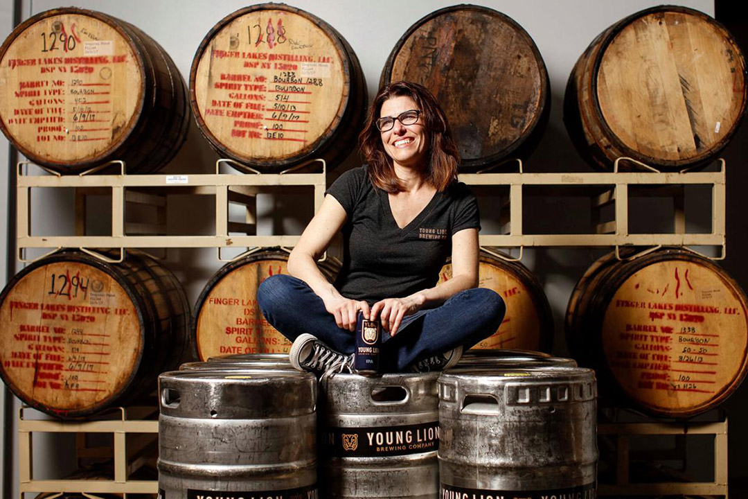 brewery owner sits on top of several kegs with stacked barrels in the background.