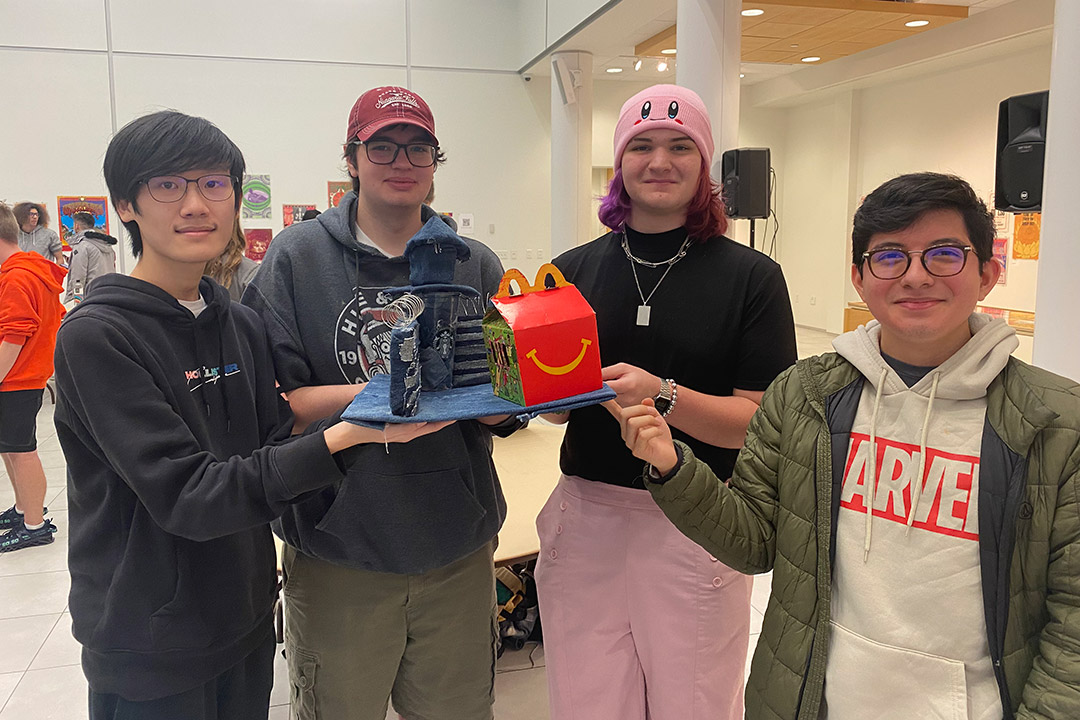 four students holding an art project depicting a jail made from jeans and a McDonalds happy meal box.