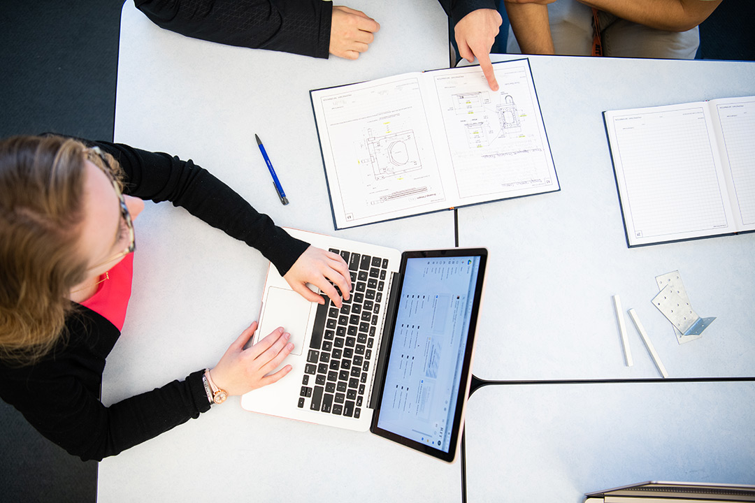 overhead view of college student working on a laptop on a table with open notebooks.