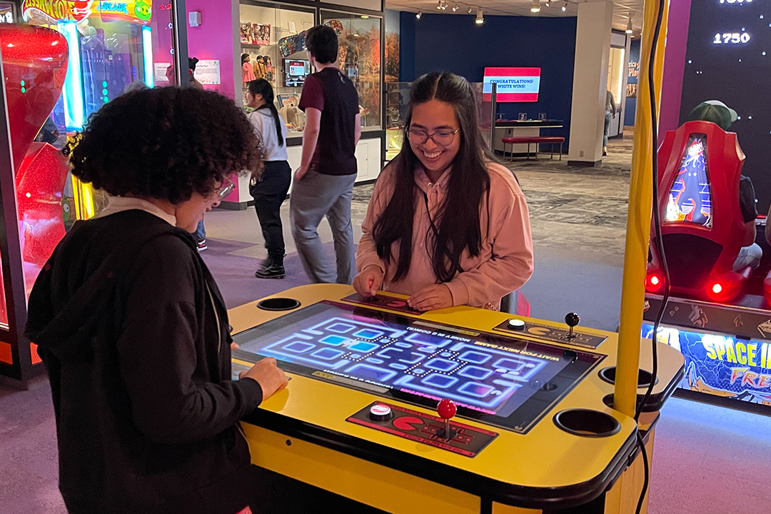 two college students playing a Pac Man game at a museum.