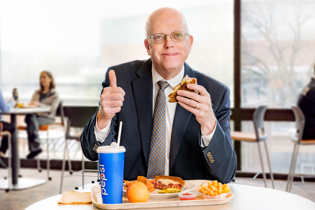 RIT President Munson sitting in a cafeteria with a plate of food in front of him, holding a sandwich in one hand and giving a thumbs up with the other.