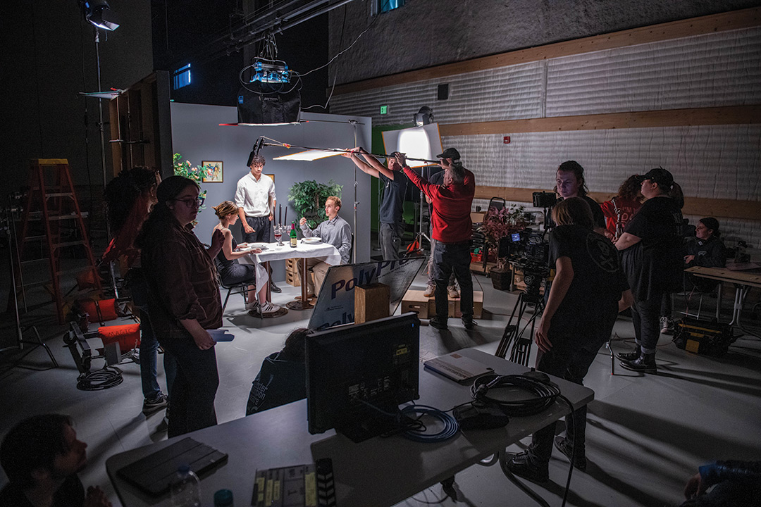 college students working on a film set with lighting, a boom mic, camera, computers, and a scene with a couple eating a meal at a table.