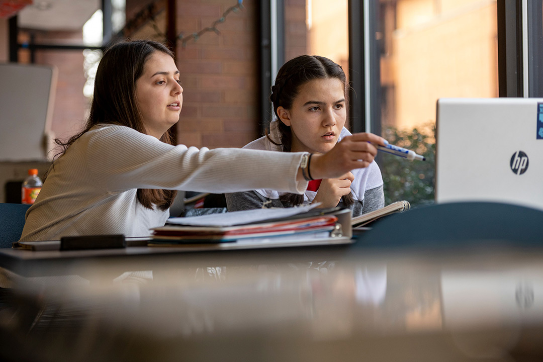 two college students sitting next to each other, with one looking on as the other points to the computer screen with a marker in her hand.