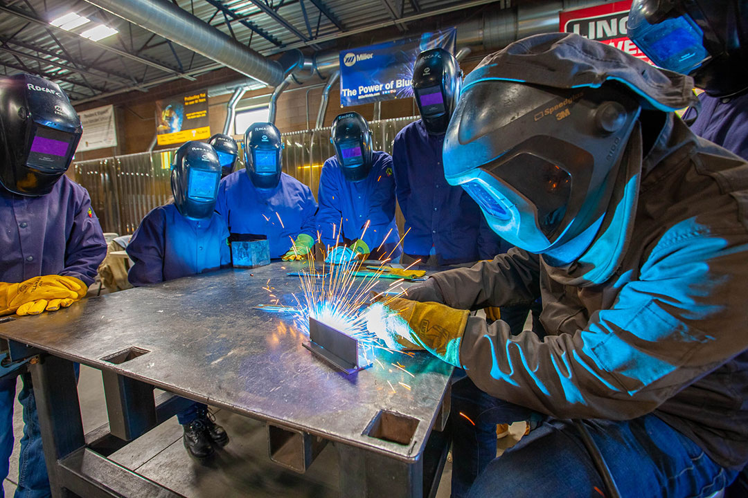 Students gather around table watching welding in blue safety suits.