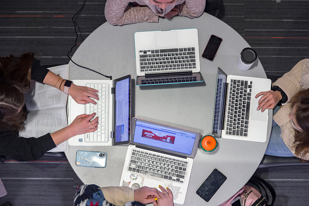 Overhead image of four students at a round table with their laptops in front of them.