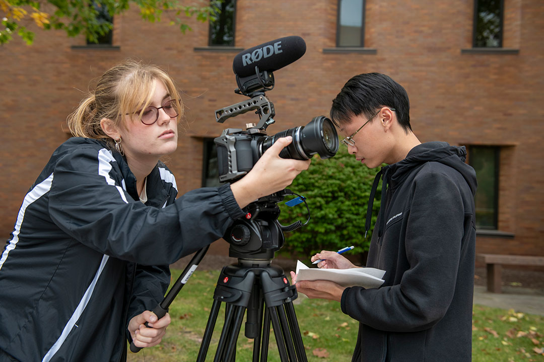 Student adjusting camera on tripod with other student writing on a sheet of paper.