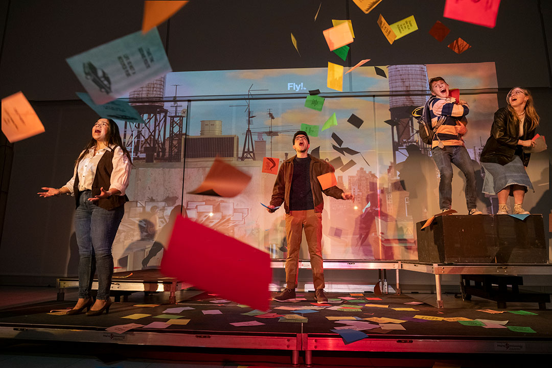 four college students on stage throwing papers into the air while rehearsing a play.