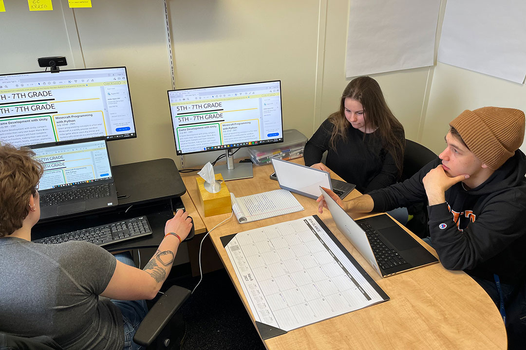 Two college students working with an RIT staff member on a desk filled with computers and paperwork