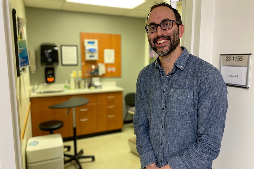 Man smiling, stands in front of a healthcare exam room.