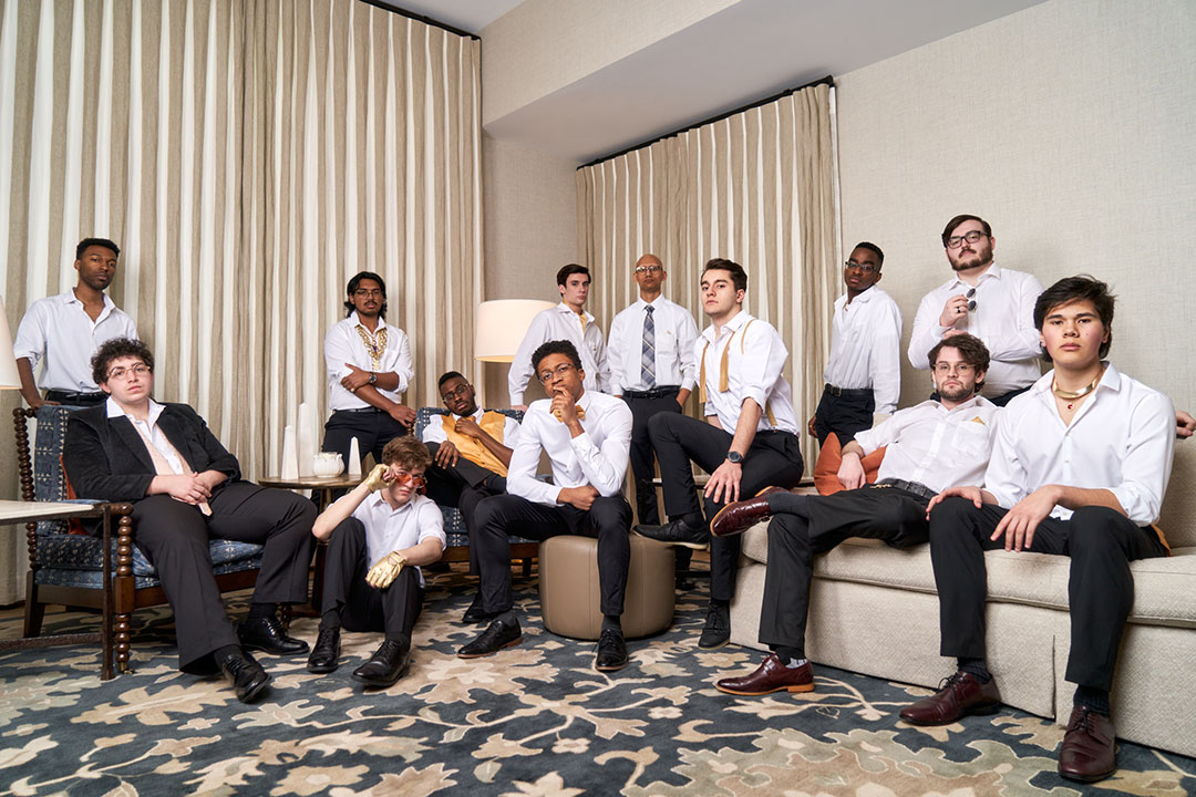 13 members of an all-male a cappella group sitting on couches and chairs,