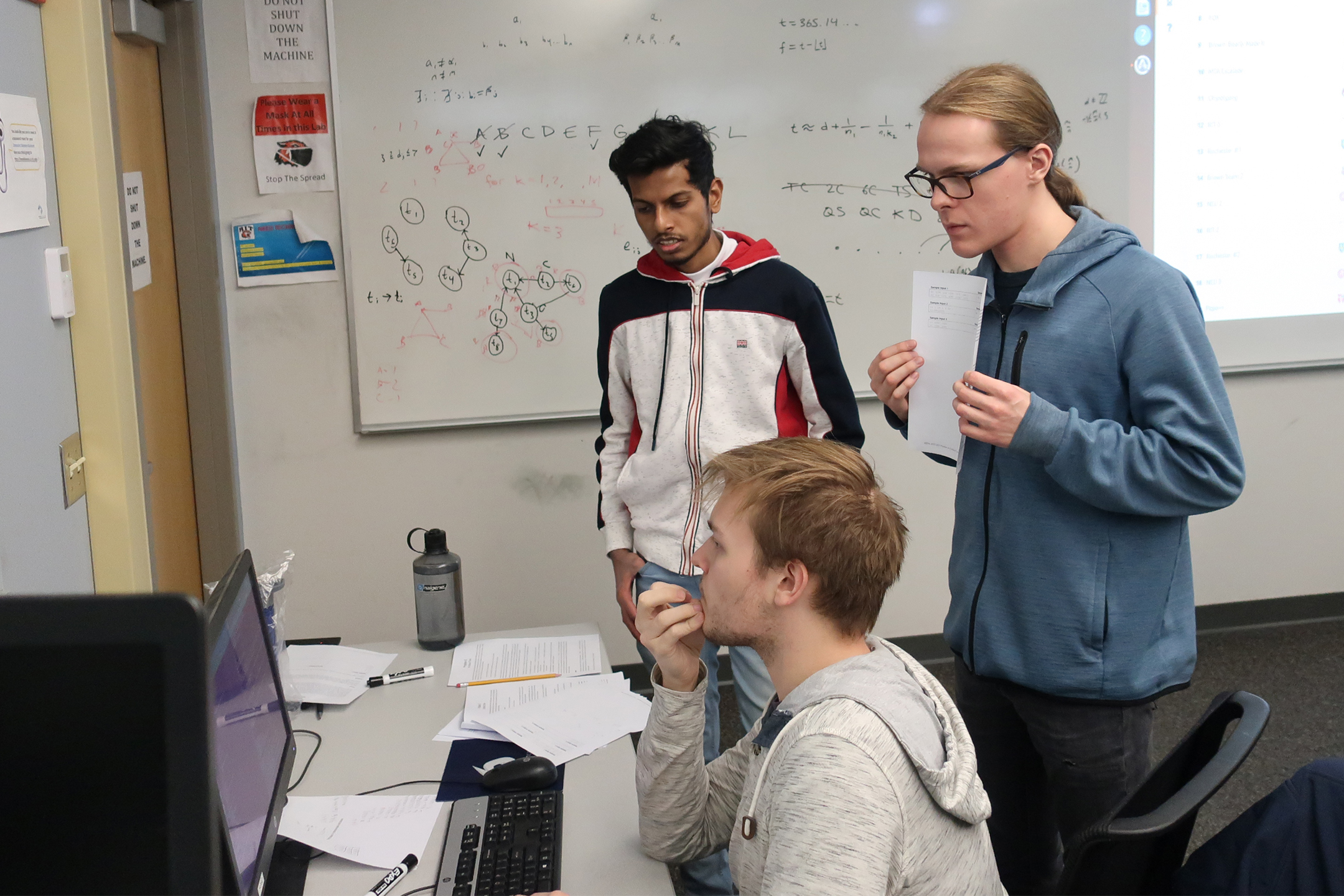 Three people stand in a classroom looking at desktop computer. 