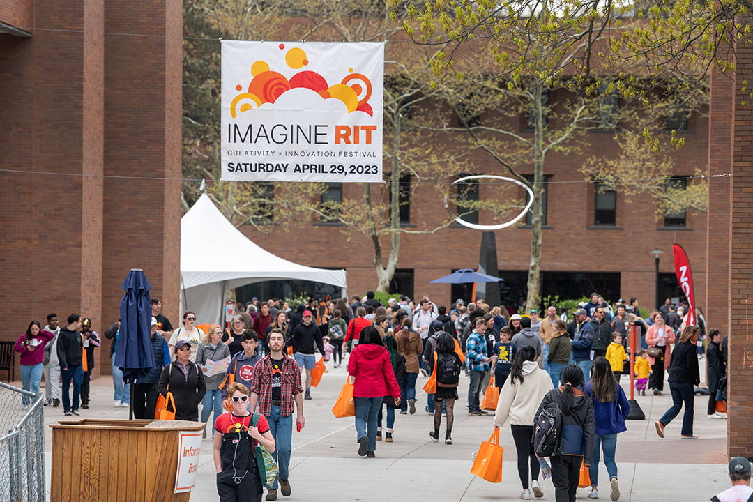 crowd of people walking in between bricks buildings under a sign for Imagine R I T.