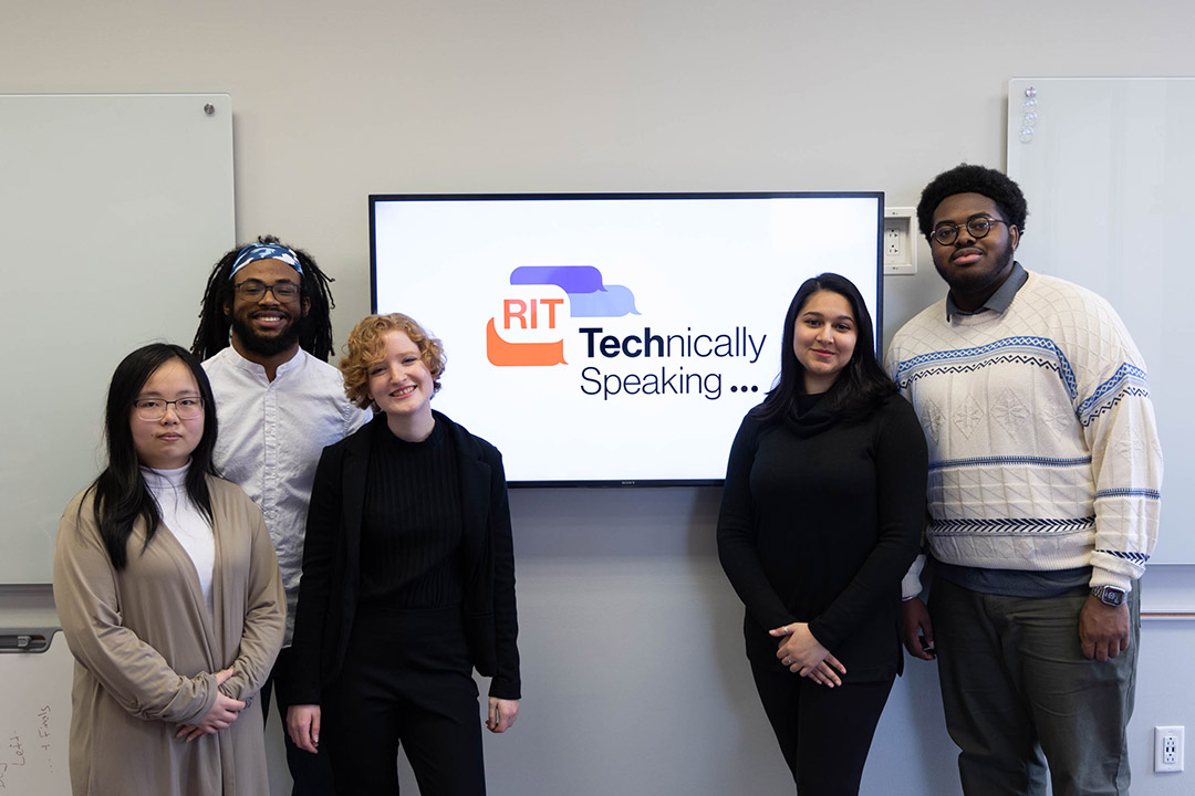 five college students standing around a TV monitor that's displaying the logo for R I T, technically speaking.