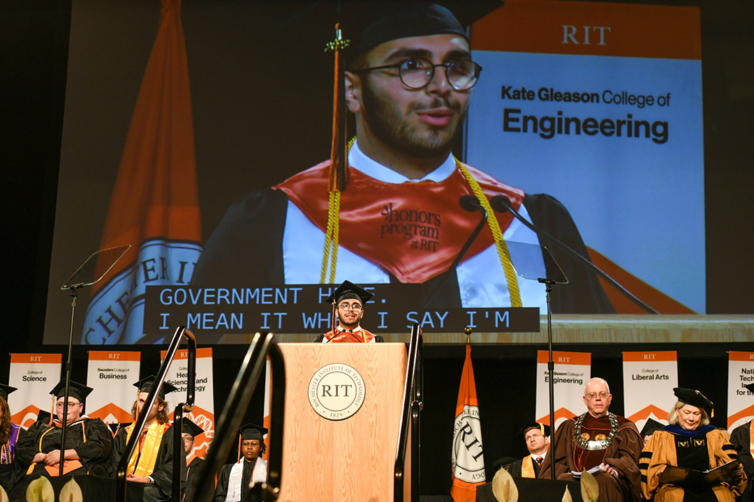 graduating student wearing regalia speaking at a podium with a giant projection behind him.