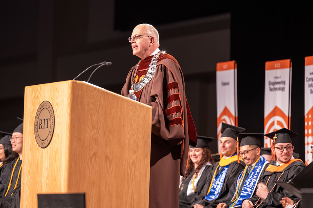 R I T president speaking at a podium wearing graduation regalia.