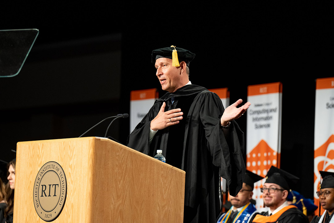 man speaking at a podium wearing graduation regalia.