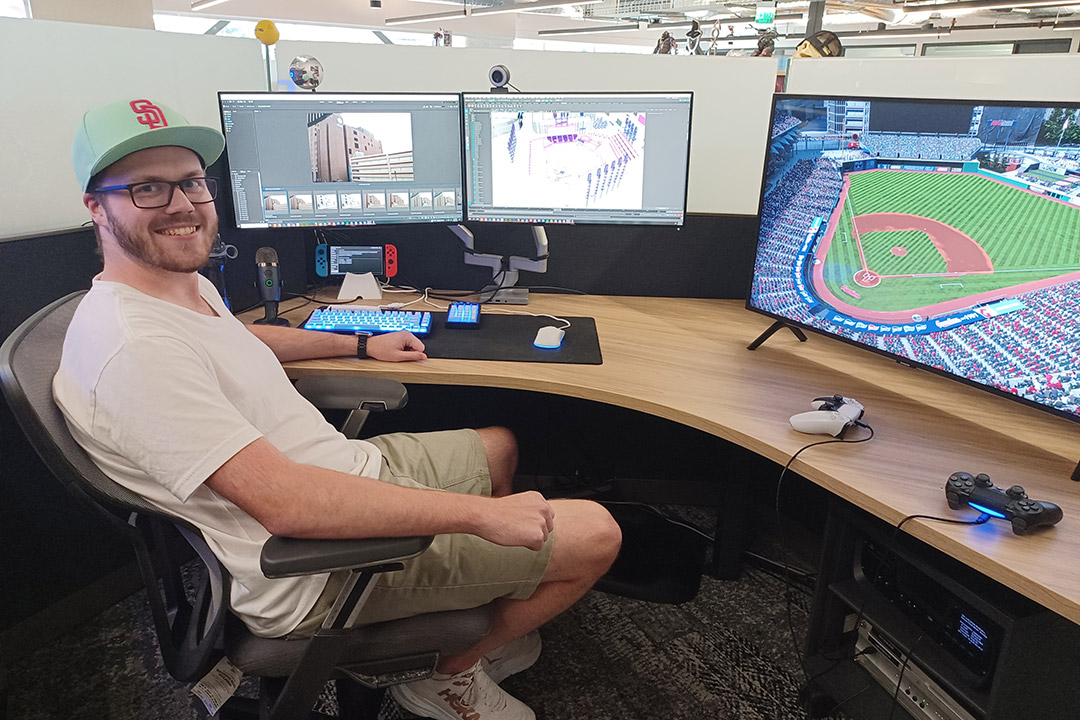 3D artist sitting at a desk with three monitors showing baseball fields and stadiums.