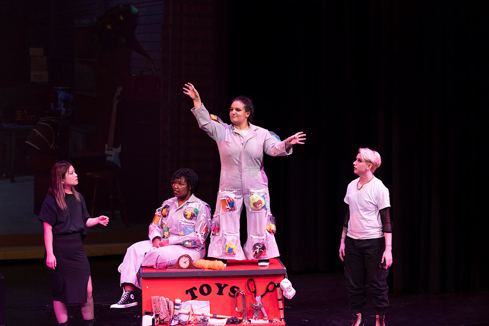 four actors on a stage, with two on top of a box labeled toys.