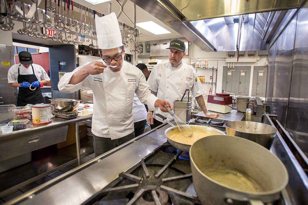 two chefs in a kitchen making sauces on a large stove, with one chef tasting a sauce with a small spoon.