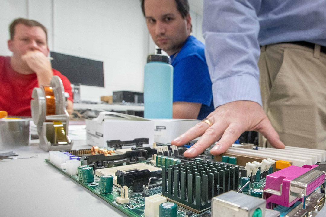 two adult student look on as a professor touches a large circuit board.