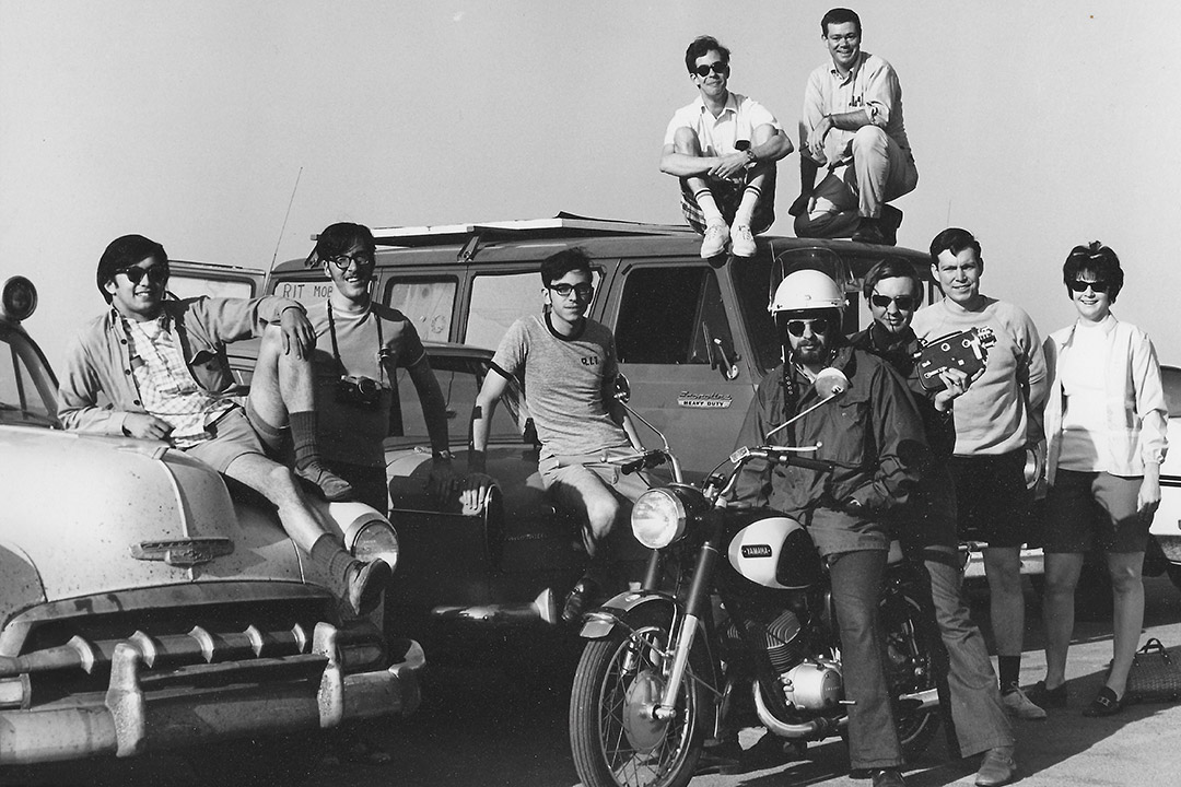 black and white photo from the 19 70s with nine college students posing on or around a car, van, and motorcycle.