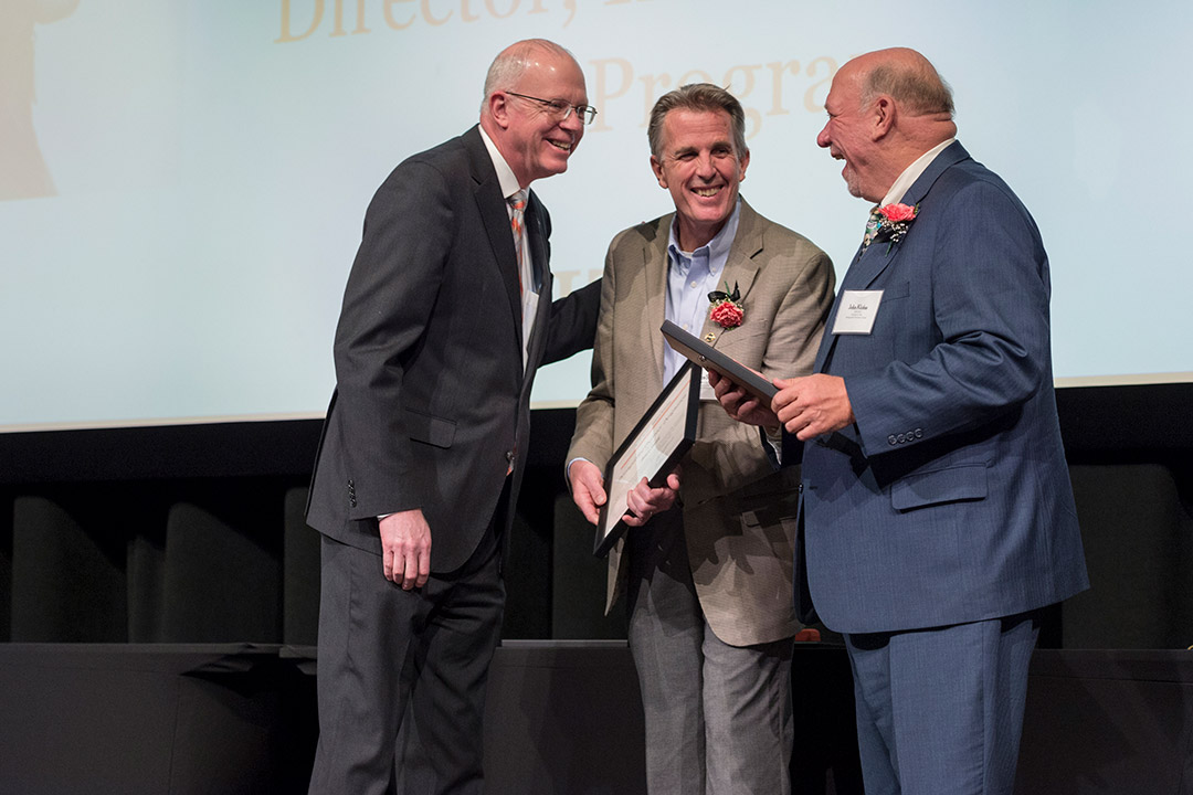 three men standing on stage, with the two on the right holding awards.