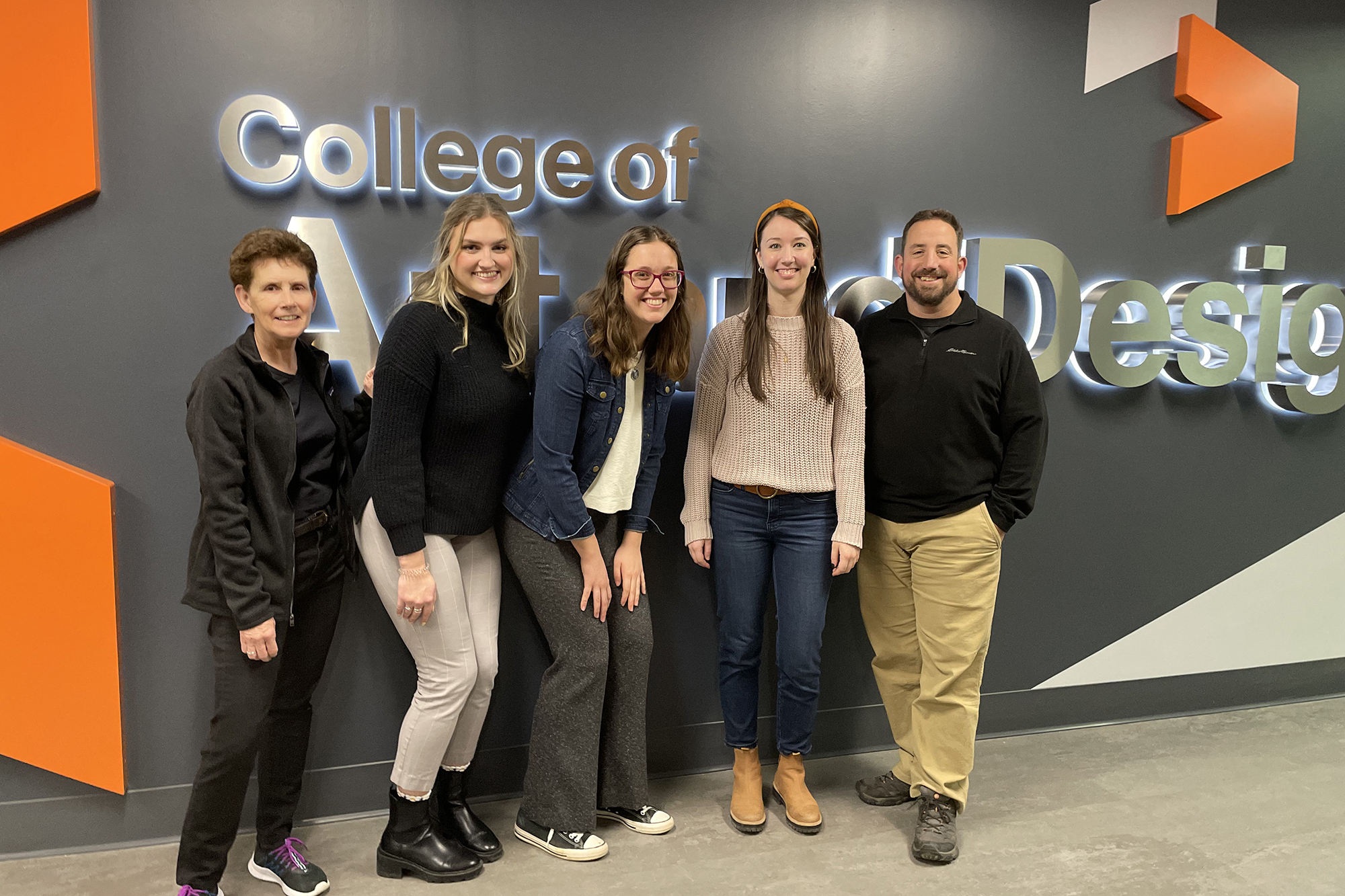 A group of alumni, faculty and industry partners pose in front of a wall sign reading College of Art and Design.